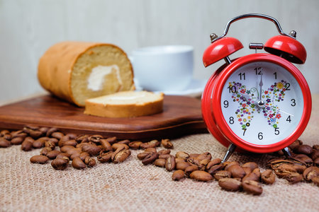 Roll coffee cake ,red alarm clock, a cup of coffee and coffee bean on wooden cutting board