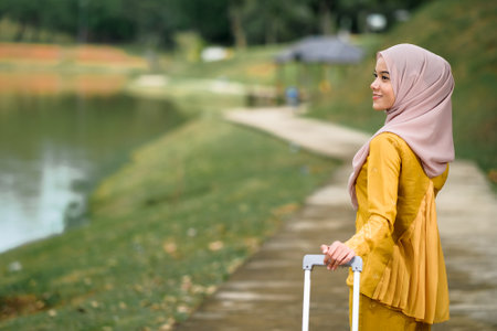 Gorgeous smiling muslim woman in traditional wear holding luggage at lake garden.の写真素材