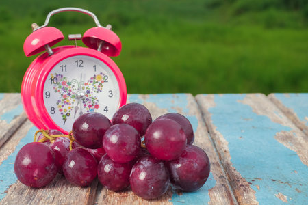 Red grapes  and alarm clock on rustic wooden with green field background , fresh fruit concept
