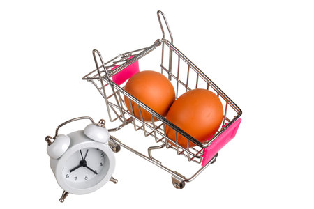 Two brown chicken eggs and alarm clock in a mini supermarket cart.Isolated on a white background.