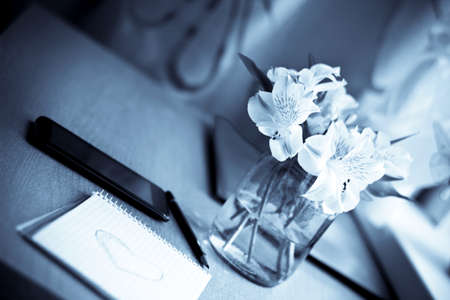 Spring composition for St. Valentines Day: bouquet of white freesia flowers, smartphone, pen and notebook with painted heart. Selective focus. Blurred multicolored background. Vintage imageの写真素材