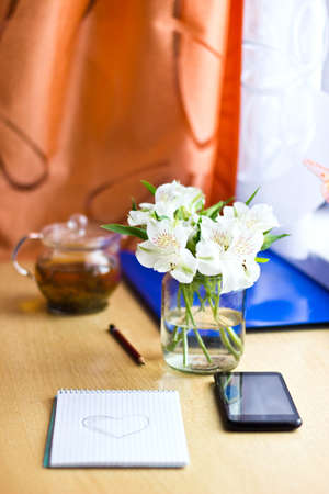 Celebrating composition: bouquet of white freesia flowers, smartphone, pen, teapot and notebook with painted heart. Selective focus. Image with multicolored bokeh backgroundの写真素材