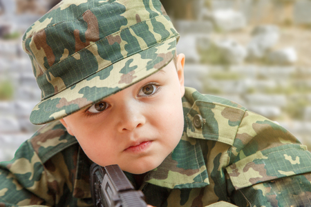 little Caucasian boy in military clothes and with toy weapons on the background of the destroyed buildingの写真素材