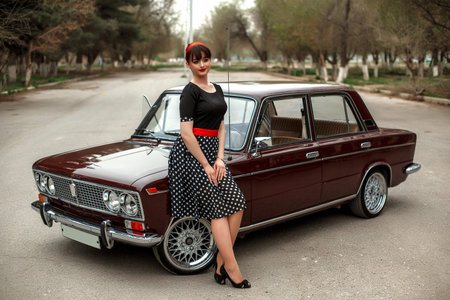 portrait of a Caucasian beautiful young girl in a black vintage dress, posing near a vintage car