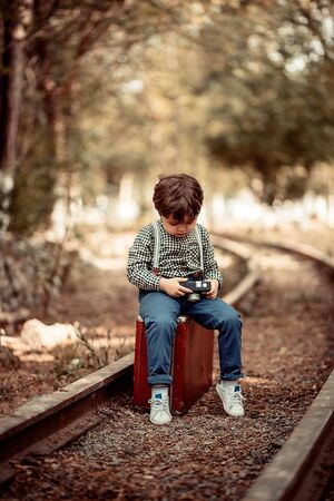 little cute boy in vintage clothes standing on an abandoned railroad with a vintage suitcase with an old camera in his handsの写真素材
