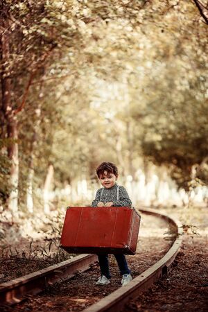 little cute boy in vintage clothes standing on an abandoned railroad with a vintage suitcase in his handsの写真素材