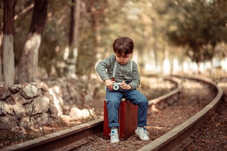 little cute boy in vintage clothes standing on an abandoned railroad with a vintage suitcase with an old camera in his handsの写真素材