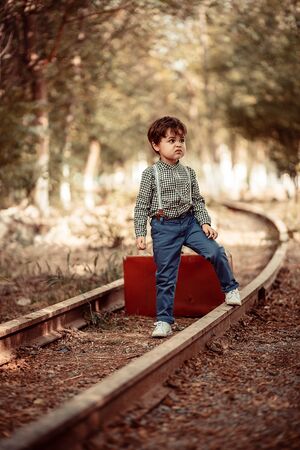 little cute boy in vintage clothes standing on an abandoned railroad with a vintage suitcase in his handsの写真素材