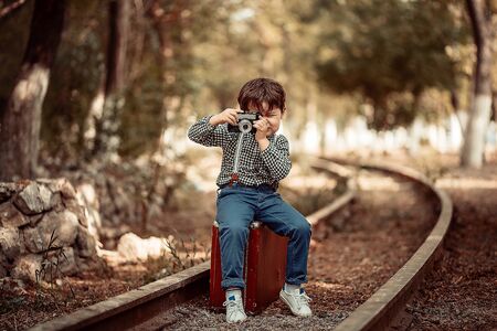 little cute boy in vintage clothes standing on an abandoned railroad with a vintage suitcase with an old camera in his handsの写真素材