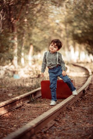 little cute boy in vintage clothes standing on an abandoned railroad with a vintage suitcase in his handsの写真素材