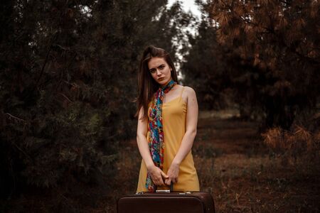 portrait of a young beautiful girl in a yellow dress with a colorful scarf standing in the woods with a vintage suitcase in handの写真素材