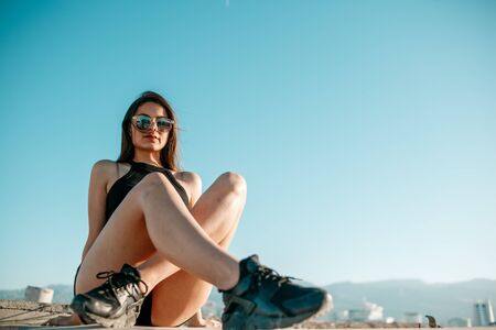 young brunette girl in black sneakers,black shorts and a black short top sitting on the roof of a tall buildingの写真素材