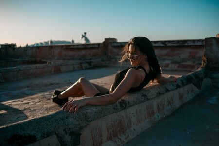 portrait of a young brunette girl in black sneakers,black shorts and a black short top sitting on the roof and enjoying the sunsetの写真素材
