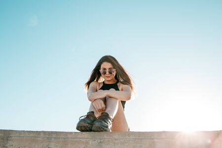 young brunette girl in black sneakers,black shorts and a black short top sitting on the roof of a tall buildingの写真素材
