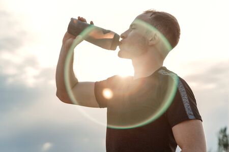 Portrait of a young Caucasian guy in a black t-shirt and black shorts drinking water from a bottle after or before trainingの写真素材