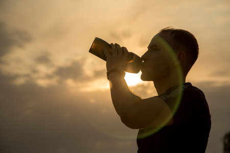 Outline of a sporty young Caucasian guy in a black t-shirt and black shorts drinking water from a...