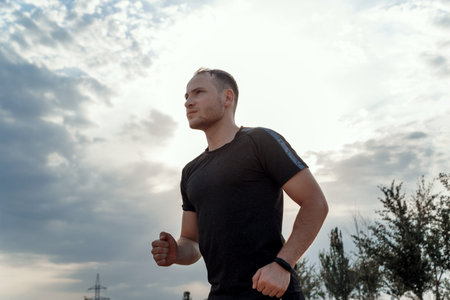 Young Caucasian guy in a black t-shirt and black shorts running over rough terrain during sunset