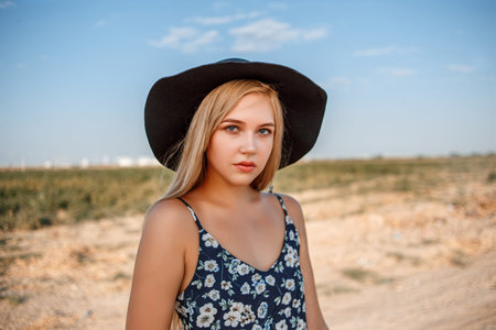 a close-up portrait of a blue-eyed blonde girl in a black hat and a floral print dress.she's posing...