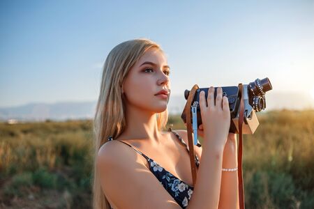 portrait of blonde girl in floral print dress with vintage video camera in grape field during sunsetの写真素材