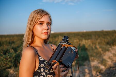 portrait of blonde girl in floral print dress with vintage video camera in grape field during sunsetの写真素材