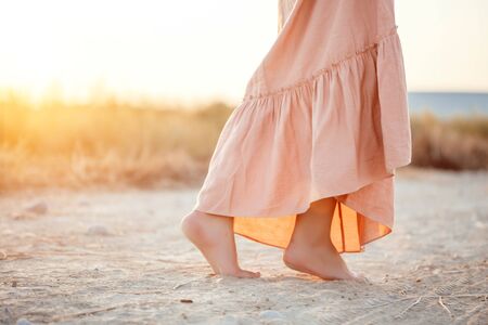 feet of a woman in a pink dress walking on the sand during sunsetの写真素材