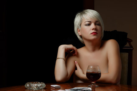 young Caucasian Nude blonde with bare Breasts sits at a table and smokes after losing at cards