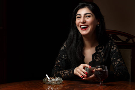 a beautiful brunette woman with scarlet lips in black clothes sits at a table and plays cards for...