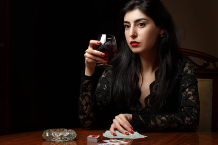 a beautiful brunette woman with scarlet lips in black clothes is sitting at a table, drinking wine and playing cards for undressingの写真素材