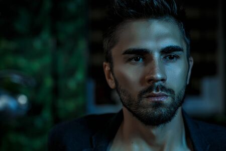 portrait of a young Caucasian guy in a black jacket on a naked body in a stylish interior under the light of neon lightsの写真素材