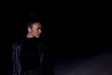 a young African American mulatto girl in a leather jacket and black clothes poses on an abandoned sand road. at night in the light of car headlights and lanternsの写真素材
