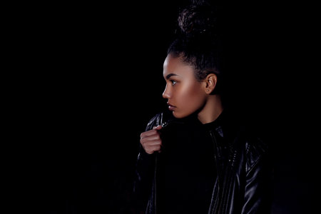 a young African American mulatto girl in a leather jacket and black clothes poses on an abandoned sand road. at night in the light of car headlights and lanternsの写真素材