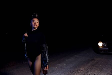 a young slender  girl in a leather jacket and bodysuit goes with a bottle of alcohol on an abandoned sandy road.at night in the light of car headlights and street lightsの写真素材