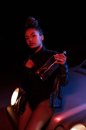 a young afro-american beautiful  girl in a leather jacket,in a bodysuit and with a bottle of...
