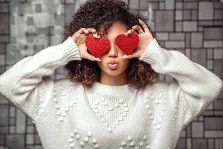 a young curly-haired African American girl in a white sweater holds two hearts in her hands . closes eyes with hearts and makes a kissの写真素材