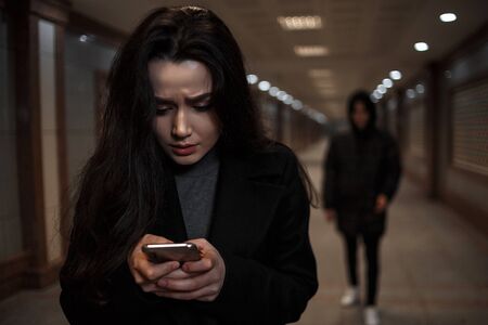 a young beautiful woman walks through an underground passage at night, followed by a man in dark clothing with a hood on his head. a woman tries to dial the number of the rescue service on the phoneの写真素材