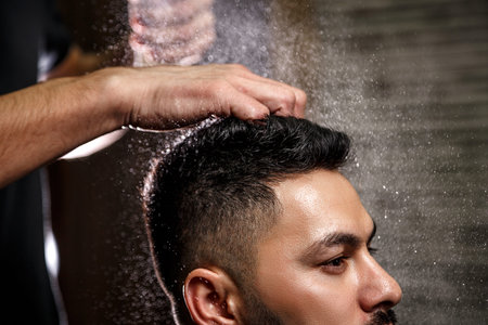 The barber dressed in a black clothes is doing the hair styling to the stylish black-haired man...