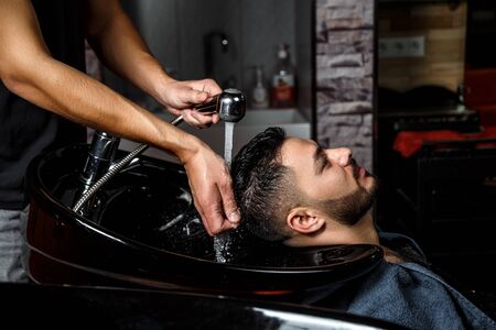a young dark-haired guy of Indian or Asian appearance in a Barber shop on a black chair. the Barber washes his head with shampoo.の写真素材