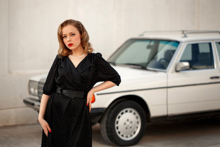 young beautiful girl in a black vintage polka dot dress next to a vintage white car.