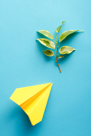 Yellow paper jet and a tree branch with green leaves on blue background.