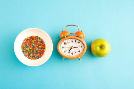 Alarm clock with a green apple and a bowl of sugar sprinkles on blue background.