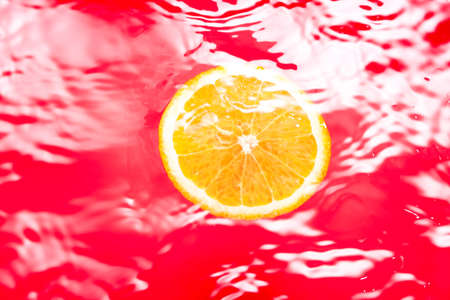 Piece of fresh orange on water surface with waves and reflections. Swimming piece of orange on red abstract background.の写真素材