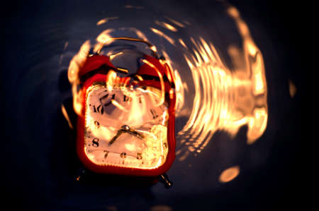 Blurry abstract composition with drowning alarm clock and ripples with fire reflected on water surface.の写真素材