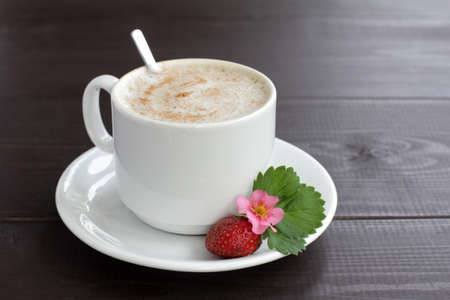 Black coffee with milk foam in white cup, decorated with strawberries, on a dark wooden table / coffee break with a strawberry aromaの写真素材