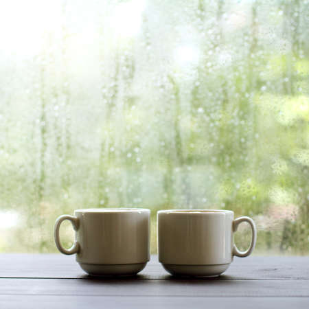 pair of white cups are on the table against the background of wet window with raindrops / warming coffee in a cozy placeの写真素材