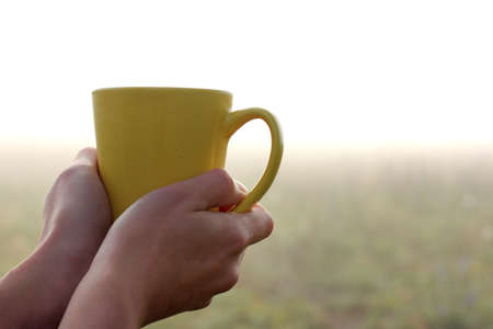 yellow ceramic mug in hand, against the backdrop the misty landscape at the beginning of the day / magic morning coffeeの写真素材