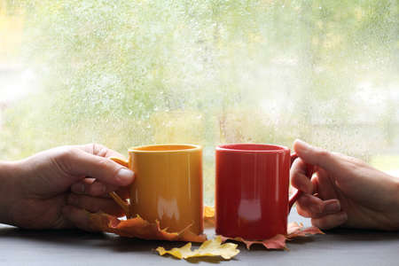 two hands holding cups on the background of wet window with raindrops / together tastier and warmerの写真素材