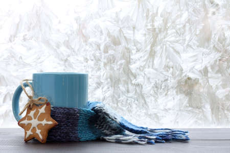 blue mug with a warm scarf and homemade cakes on the background of frozen window / drinks for the winter seasonの写真素材