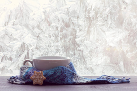 White empty mug for warming beverage wrapped in a scarf on a table next to a frosty winter window /...