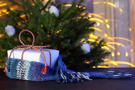 gift wrapped in a woolen scarf lying under the tree, on a background of festive lights flying in the background / warming Christmas atmosphereの写真素材