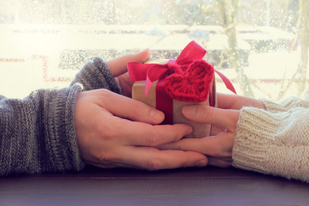 couple holds in hands gift with a heart symbol against the window / holiday greeting with a hint